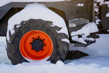 Snow covered heavy equipment wheel with bright orange rim and thick tire tread, industrial vehicle step and chassis detail in winter yard on frozen ground