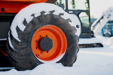 Snow covered heavy equipment wheel with bright orange rim and rugged tire tread, parked industrial machine in winter yard with blurred lift vehicle in background