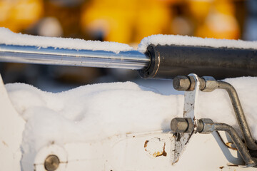 Snow resting on chrome hydraulic piston rod and black seal housing, macro detail of heavy machinery bracket bolts and steel lines in cold winter light