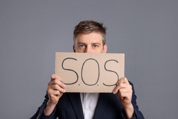 Unhappy businessman holding cardboard sign with word SOS on grey background