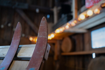 Two old ski tips and binding edge in focus, warm fairy lights bokeh and wooden interior behind, rustic storage scene for winter outdoor sports gear