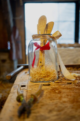 Handmade gift in glass jar with wooden angel figures, red ribbon bow and wood shavings on carpentry workbench, rustic workshop background with window