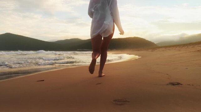 Close-up of woman legs in white shirt standing barefoot on wet sand and run in sunset. Gentle waves, golden sky. Peaceful and tranquil setting, reflecting calmness, grounding and nature connection