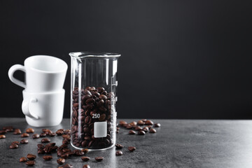 Coffee beans in glass beaker and cups on grey table, closeup. Space for text
