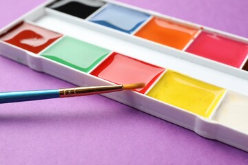 Palette with watercolor paints and brush on purple background, closeup
