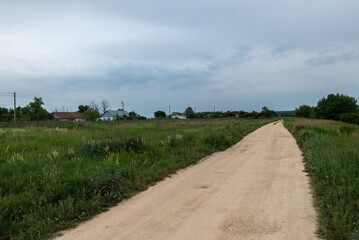 Rural dirt road leading to distant village under cloudy sky