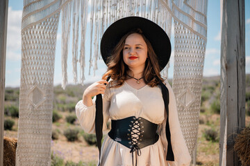 Woman stands in a rural setting wearing a hat and posing near decorative fabric in sunlight