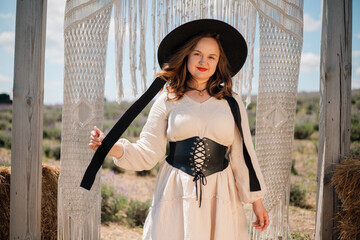 Woman stands in a rural setting wearing a hat and posing near decorative fabric in sunlight