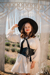 Young woman stands in outdoor setting wearing hat and dress while holding a bow of fabric, surrounded by natural scenery in sunny weather