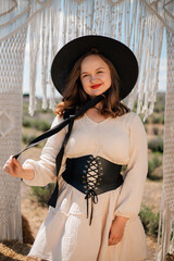 Young woman stands in outdoor setting wearing hat and dress while holding a bow of fabric, surrounded by natural scenery in sunny weather