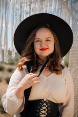 Young woman stands in outdoor setting wearing hat and dress while holding a bow of fabric, surrounded by natural scenery in sunny weather