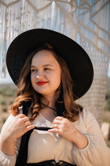 Young woman stands in outdoor setting wearing hat and dress while holding a bow of fabric, surrounded by natural scenery in sunny weather