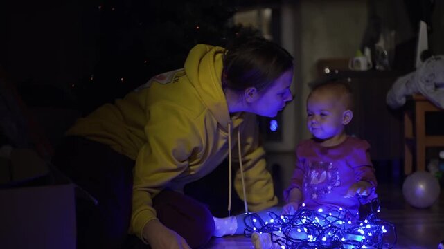 mother kisses her baby daughter, they cheerfully raise their hands up and play, bump their foreheads, look at the bright blue garland, the mother lies down on her daughter s feet