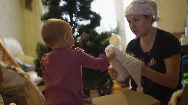 mother persuades baby girl put bright shiny white New Year's hat like hers child quickly takes head asks put, then points finge camera. background Christmas tree waiting decoration with toys and