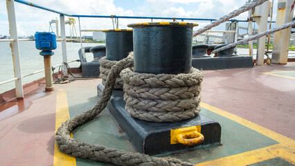 Close-up of a heavy mooring bollard with thick nautical rope on a ship deck. Industrial port background with cranes under a cloudy sky. Nautical maritime concept