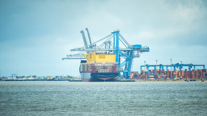 Large container ship docked at a busy industrial port with blue gantry cranes, cargo containers, and a cloudy sky. Perfect for logistics and trade