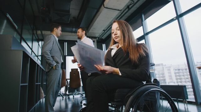 A group of professionals discusses ideas about inclusion and diversity in a busy coworking office. One woman uses a wheelchair while reviewing documents.