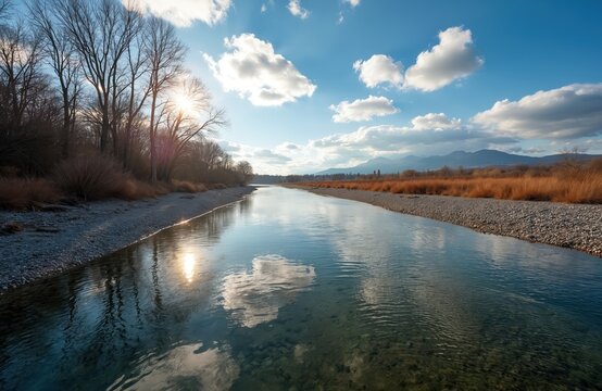 Wide river flows through dry grass banks under a blue sky with fluffy clouds. Bare trees line the shore, sun reflects on water surface. Distant mountains are visible on horizon.