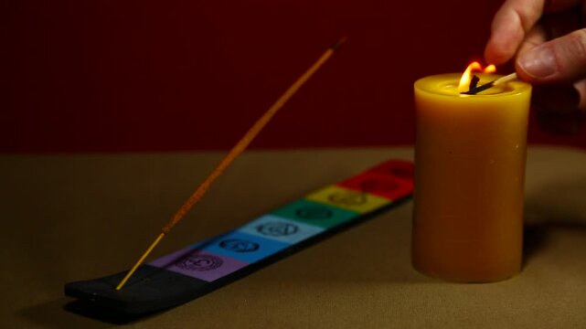 Woman hand lights the candle fire near a smoldering aroma incense on decor incense holder with colorful colors symbols of chakras on red background. Yoga therapy, spa fumigation