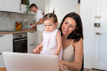 Smiling mother works on a laptop with her baby daughter on her lap while partner cooks in the kitchen, balancing remote work, childcare and everyday family life at home