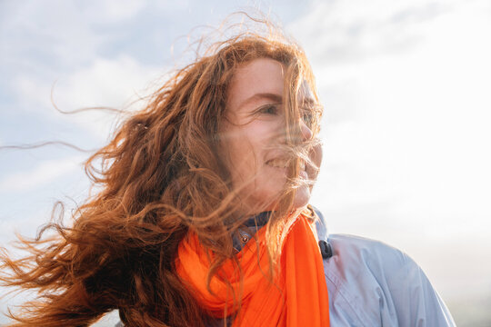 Redhead Woman with long hair smiling against cloudy sky while wearing orange scarf at outdoor location