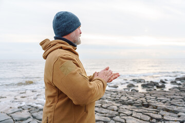 Man stands by the shore looking at the water as clouds drift above during daytime in cold weather © Iryna