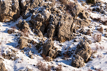 Large grey boulders and jagged rock formations are covered in white snow on a steep mountain side under the soft light of a cold winter day in the wild