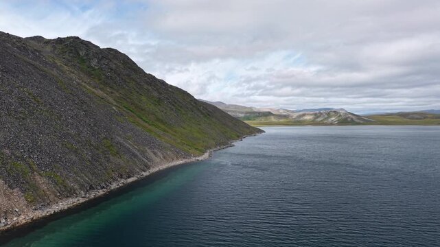 Aerial view of a high rocky slope above a narrow green water edge in Russia, Chukotka, Lavrentiya Bay at daytime under broken cloud. Minimal wake and clear color serve premium travel need
