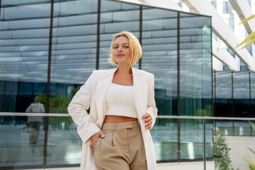 Young businesswoman with short blond hair in blazer posing near modern glass building with successful and professional appearance in city