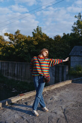 Fototapeta premium Woman in a colorful sweater and jeans posing outdoors on a rural rooftop with a crossbody bag, playful pose in warm sunlight for casual fashion, lifestyle portrait and seasonal street style.