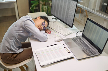 Woman employee in office tired, sleeping. She naps at a desk with laptop and monitors. Overworked by deadlines and data entry. Paperwork unfinished on the desk. Work fatigue and stress concept.