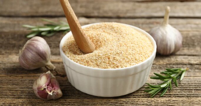 Woman stirring granulated dried garlic with spoon, rosemary and fresh vegetables at wooden table, closeup