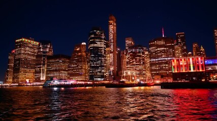 City lights shine brightly in New York at night. The water reflects the colorful buildings. People enjoy the lively ambiance. Boats move through the water creating ripples.