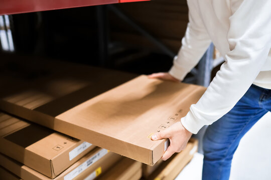 Close-up of a person pulling a flat-pack cardboard box from a shelf in a large warehouse or self-service furniture store. Concept of logistics, shopping, and home improvement