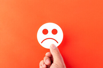 A hand holding a white paper cutout of a sad face emoji over an orange background. Perfect for concepts of customer dissatisfaction, negative feedback, mental health, and disappointment.