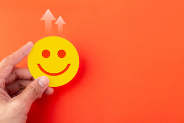 A hand holding a yellow happy face emoji icon on an orange background with upward arrows. Concept for business growth, customer satisfaction, positive feedback, and financial success.