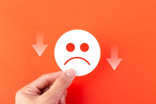 A hand holding a white sad face emoji icon on an orange background with downward arrows. Concept for business failure, customer dissatisfaction, negative feedback, and economic decline.