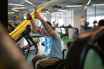 Concentrated disabled man in wheelchair working on spinal injury rehabilitation at gym, doing sports exercise for strength in upper body, using fitness machine for power workout on daily session.