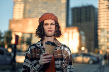 Young person holding coffee on urban street, beanie and plaid jacket, skeptical expression captured...