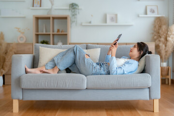 Happy cheerful positive young Asian woman on the phone at home, smiling girl using app and browsing on the internet on smartphone lying on sofa