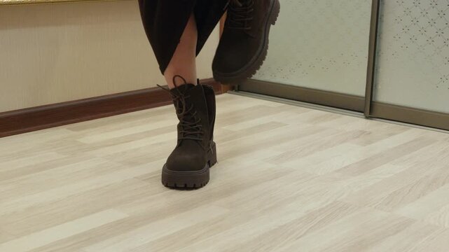 Adult woman taking off dark boots indoors, close up of feet and hands on wooden floor. Home routine, comfort and relaxation concept.