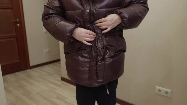 Adult woman standing indoors fastening brown winter jacket with hands, preparing to go outside. Cold season clothing, daily routine and lifestyle concept.