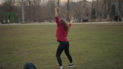 Lady Jumps On Green Grass, Female Athlete In City Park Performing Stunt, Young Woman Dressed In Red Hoodie Balancing Confidently On Grassy Area With Cityscape Behind And Calm Sky Above