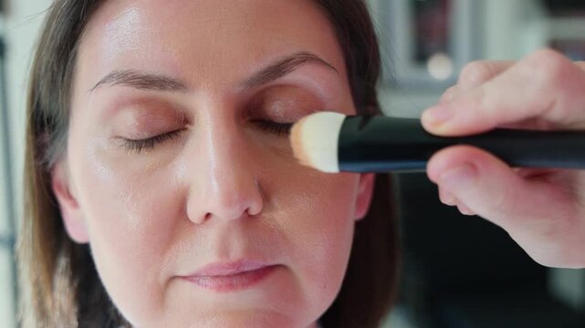 Closeup of a mature woman with closed eyes while a makeup artist applies liquid foundation with a brush in a professional beauty salon. Skincare, cosmetics, wellness, pampering concept