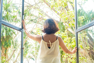 The woman was at the window in the bedroom. She opened the curtains on the window. In the morning and she looks at the view of mountains and trees at sunrise.	