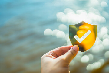 Hand holding golden shield with check mark over blurred water background, symbolizing security, protection, trust, safety, insurance assurance, risk prevention, and reliable protection concept.