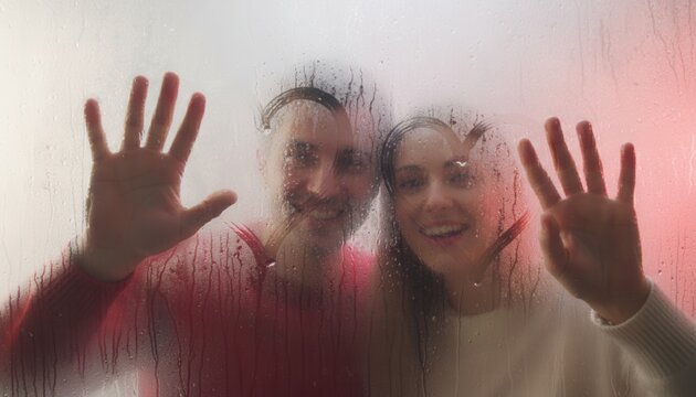 A smiling couple behind wet, steamy glass, pressing hands and showing a drawn heart shape, Valentine's Day concept.