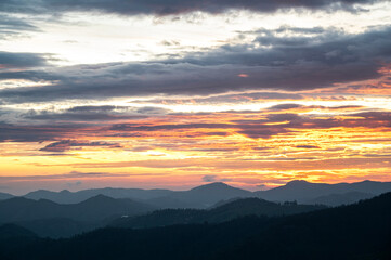 Landscape sunrise near Ooty, Nilgiris showing layered mountain silhouettes emerging through mist on a monsoon morning, with a cloudy sky glowing in soft yellow, orange, black and pink gradients.