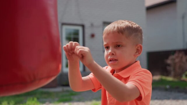 Child Strikes Red Bag Happily, Young Boy Enthusiastically Punches Bright Red Bag In Sunlight, Energetic Caucasian Boy Delivers Playful Punches To Colorful Bag Outdoors In Sunny Yard