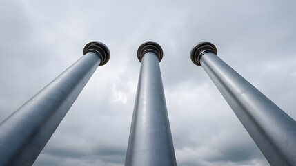 Three tall metallic columns ascend towards an overcast sky from a low angle perspective
